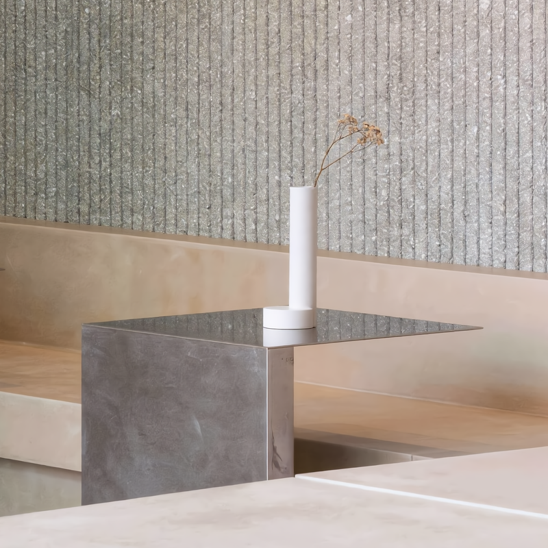 Minimalist tall cylindrical plaster vase styled on a raw industrial brushed steel side table, showcasing luxury brutalist design aesthetics for interior architecture mood boards.